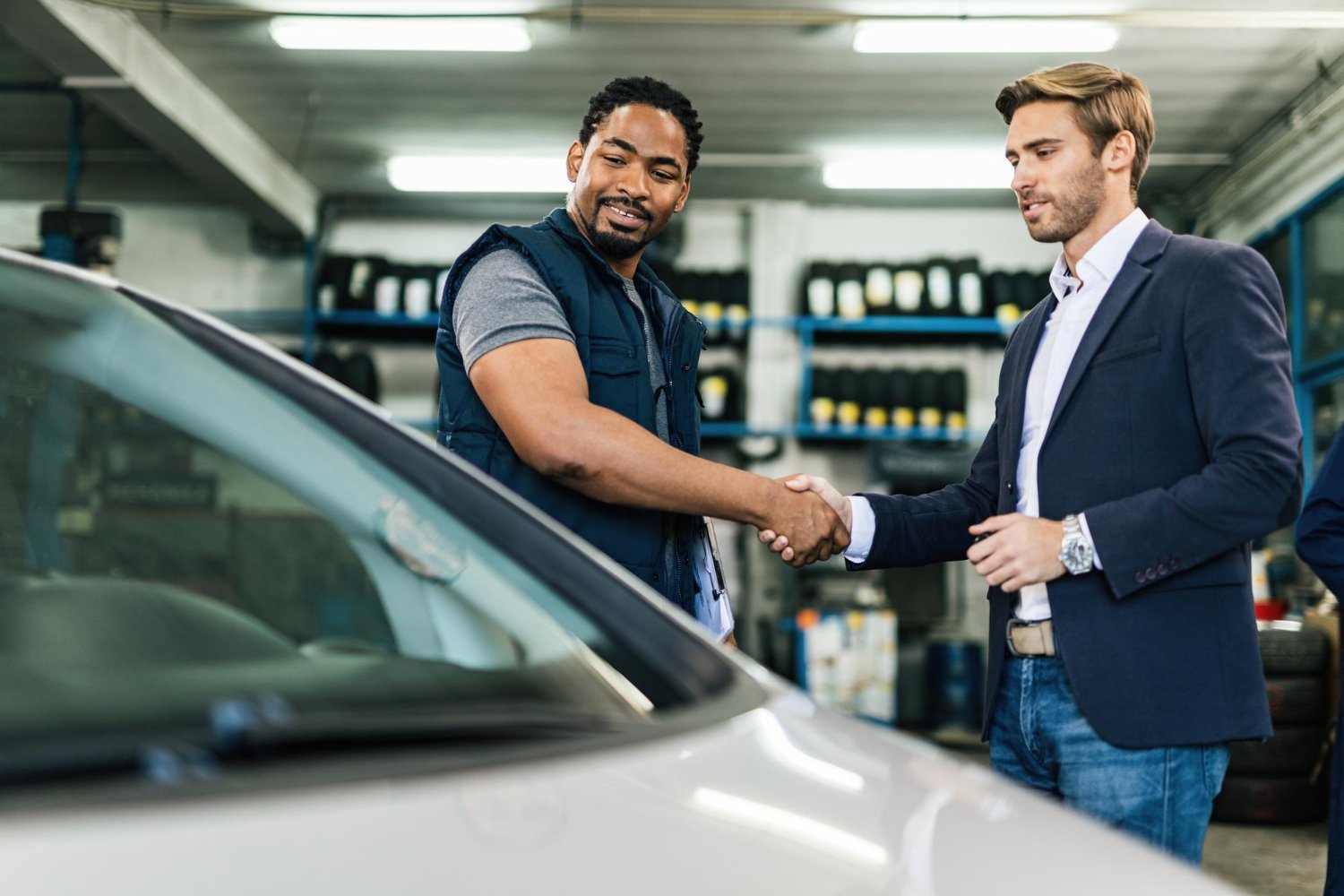 dans atelier en font des pneu ranger en premier plant on vois 2 hommes se serrant la main devant une voiture